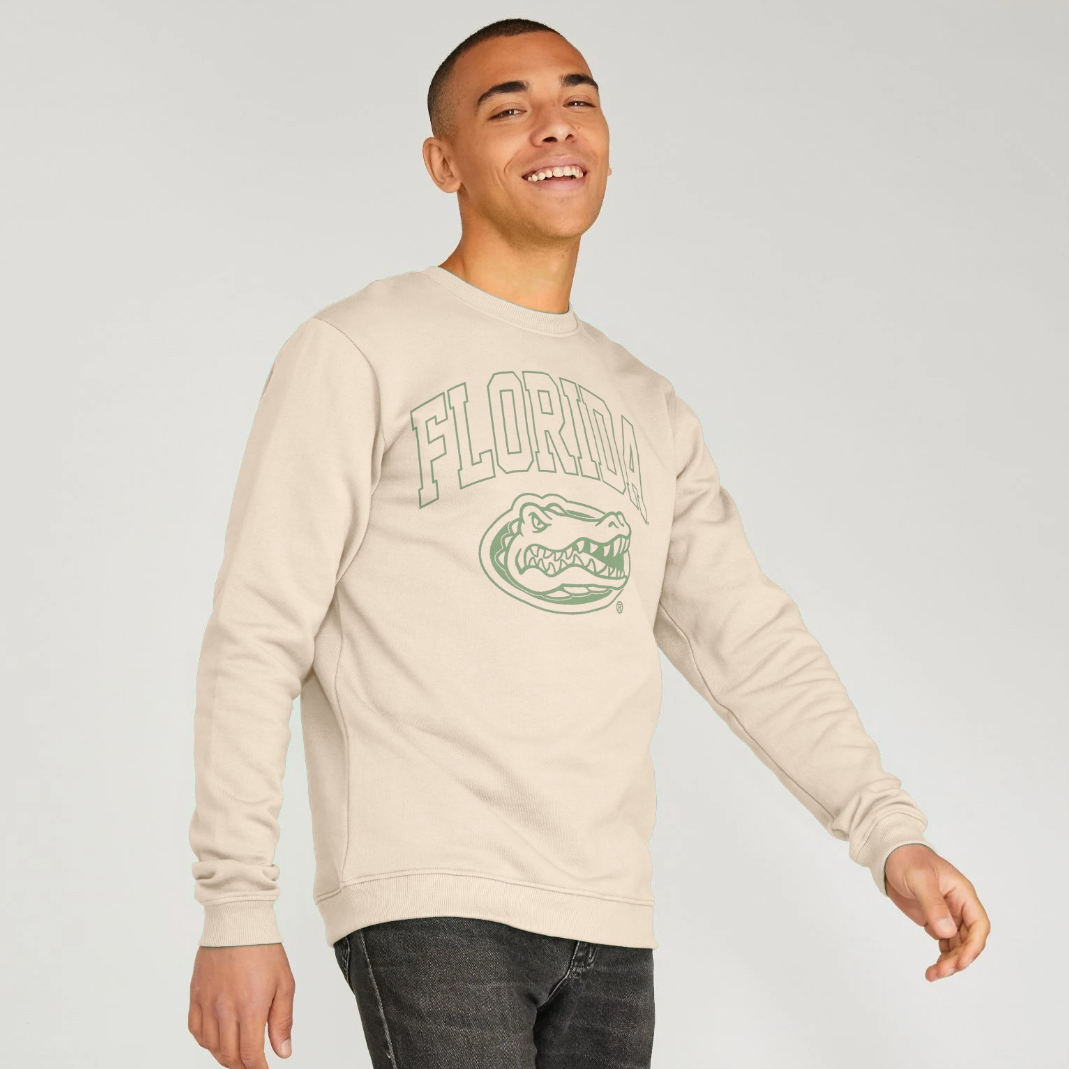 A man wearing a cream-colored crewneck sweatshirt with a green Florida Gators graphic on the front, paired with dark gray jeans, against a light gray studio background.