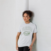 A woman with curly hair leans against a white brick wall, wearing a white t-shirt with a green Florida Gators graphic on the front.