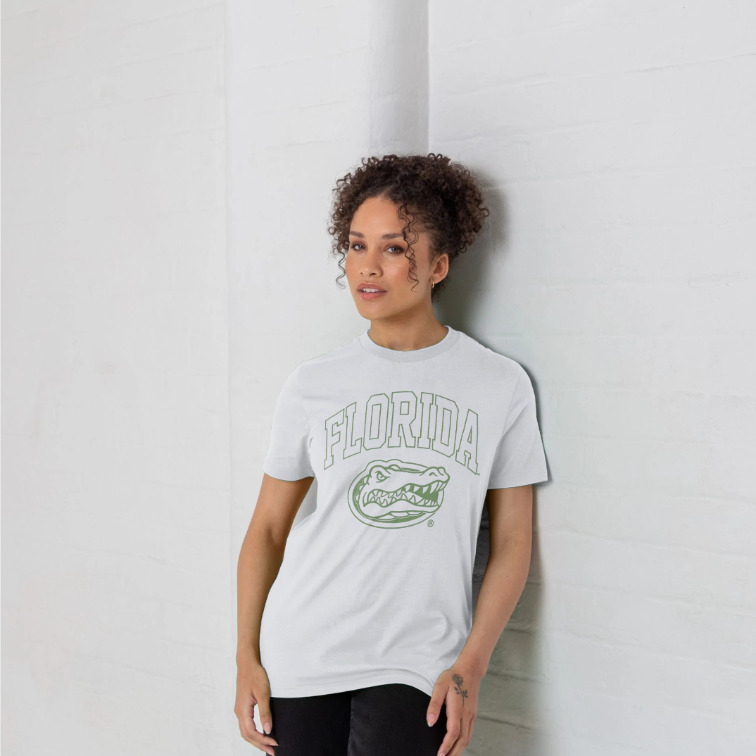A woman with curly hair leans against a white brick wall, wearing a white t-shirt with a green Florida Gators graphic on the front.