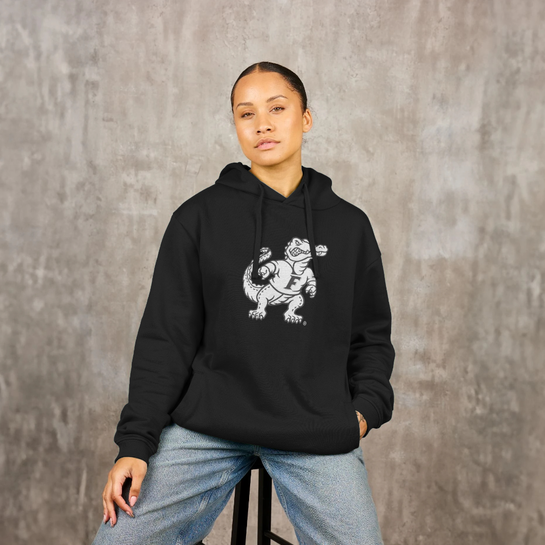 A woman wearing a black pullover hoodie with a white graphic of a Florida Gator on the front, paired with light blue jeans, sitting on a stool against a textured gray wall.