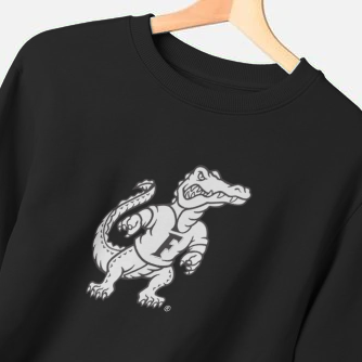 A black crewneck sweatshirt hanging on a wooden hanger against a light gray background, featuring a white graphic of an alligator wearing a football uniform on the chest.