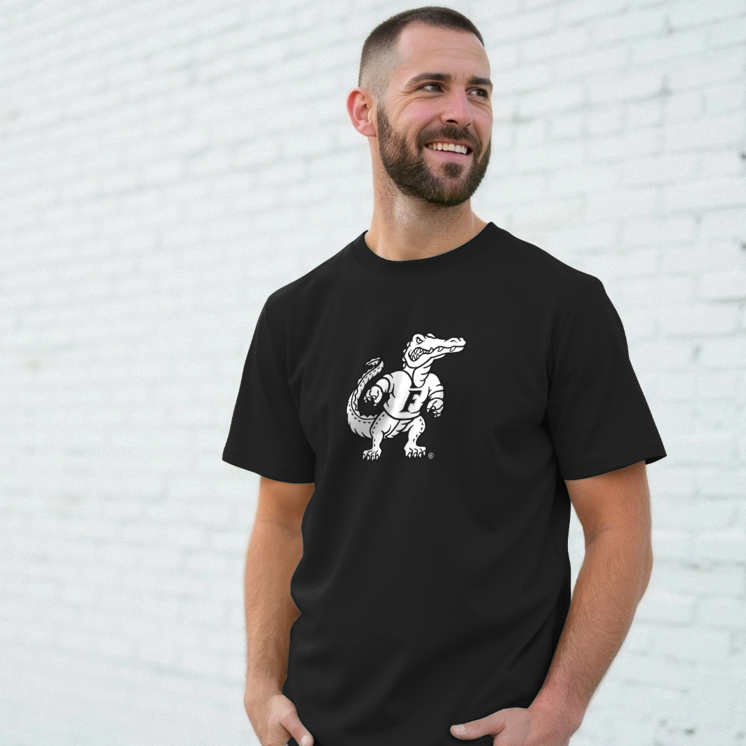 A man wearing a black crewneck t-shirt with a white graphic of a cartoon alligator  standing against a white brick wall.