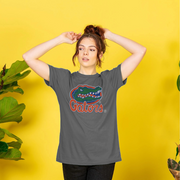 A woman wearing a dark gray t-shirt with a Florida Gators logo graphic, posing against a bright yellow background with decorative plants.