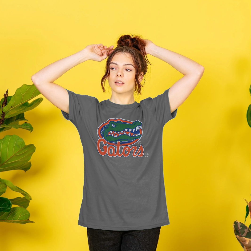 A woman wearing a dark gray t-shirt with a Florida Gators logo graphic, posing against a bright yellow background with decorative plants.